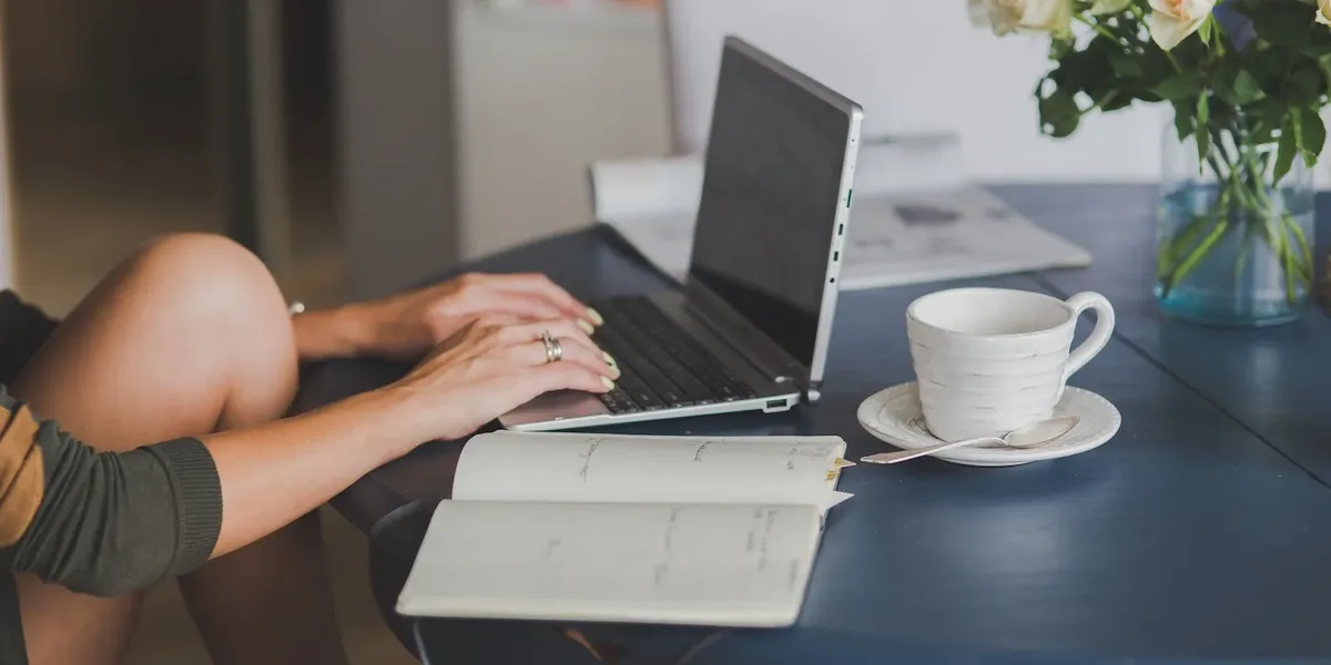 Hands typing on a laptop beside a notebook of ideas and a cup of tea