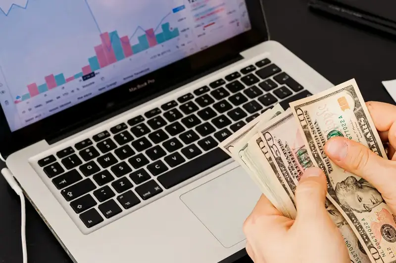 Hands counting US dollar bills in front of a laptop showing a growth chart