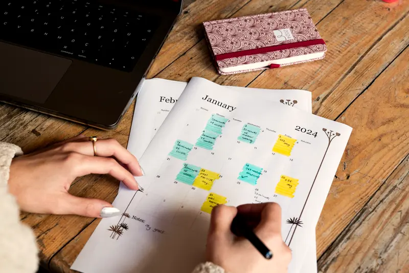 Hands marking a paper calendar planner with sticky notes on a wooden desk