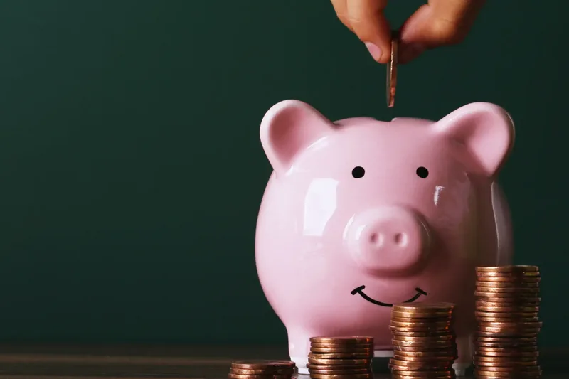A hand dropping a coin into a pink piggy bank beside growing stacks of coins