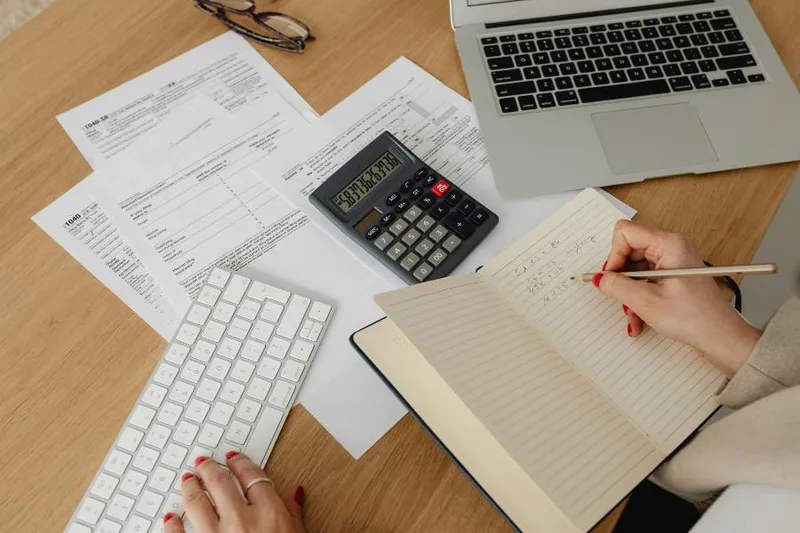 A desk with income paperwork, a calculator, an open notebook and a laptop while budgeting
