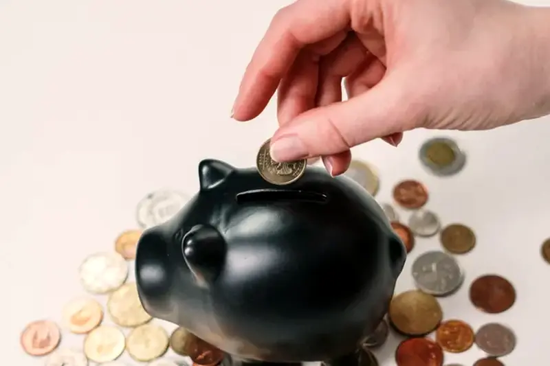 A hand putting a coin into a black piggy bank surrounded by coins