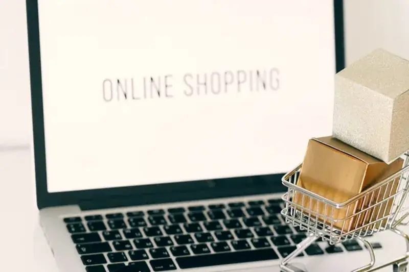 A laptop showing online shopping with a miniature cart and delivery boxes
